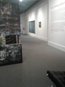 section of AGN gallery with free standing wall on left side of image. You can see a window in the wall and in the background some two dimensional art is visible