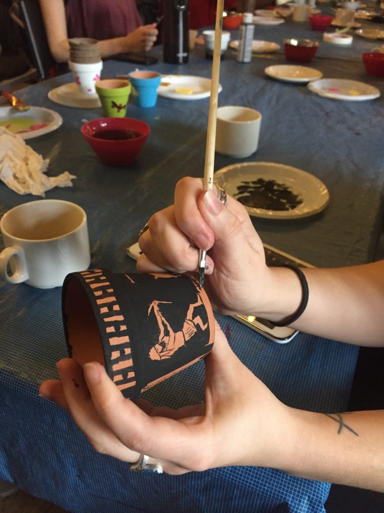 Two hands holding a small 2 inch terracotta pot and paintbrush. Painting the pot to look like Greek Black-Figure art. In the background people are painting similar sized pots in bright colours and patterns as part of a Social Prescribing event.